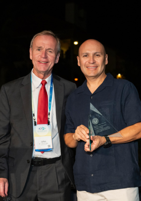 A photo of a man accepting the ACP Award of Distinction: he stands on stage, smiling proudly, holding the elegant award plaque as the audience applauds in the background, with a banner or screen displaying the event logo and accolades.